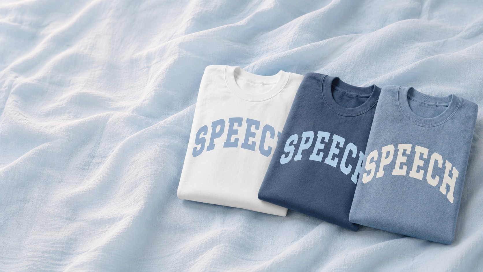 Three folded SPEECH crewnecks in white, denim, and blue jean on sunlit linen